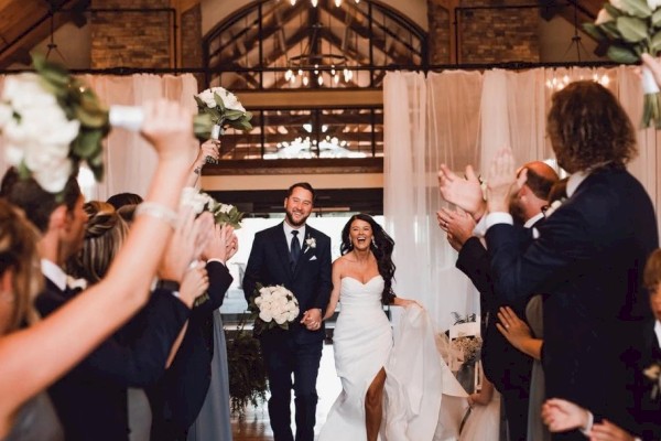 A newlywed couple walks down an aisle of cheering guests in a rustic wooden chapel, wedding party and guests applauding as they celebrate.