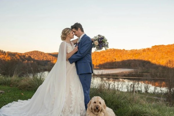 A newlywed couple shares a kiss by a lakeside, with a fluffy dog lounging nearby as golden light bathes the scene at sunset.