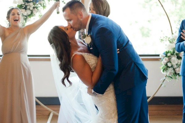 A newlywed couple shares a kiss while dancing; the bride in a white gown and the groom in a blue suit, with a bridesmaid cheering in a beige dress nearby.