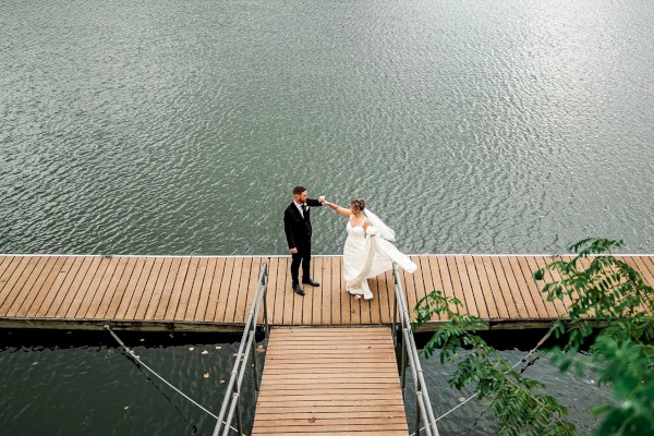 A couple in wedding attire dances on a wooden dock extending into calm water, holding hands as they spin under soft daylight.