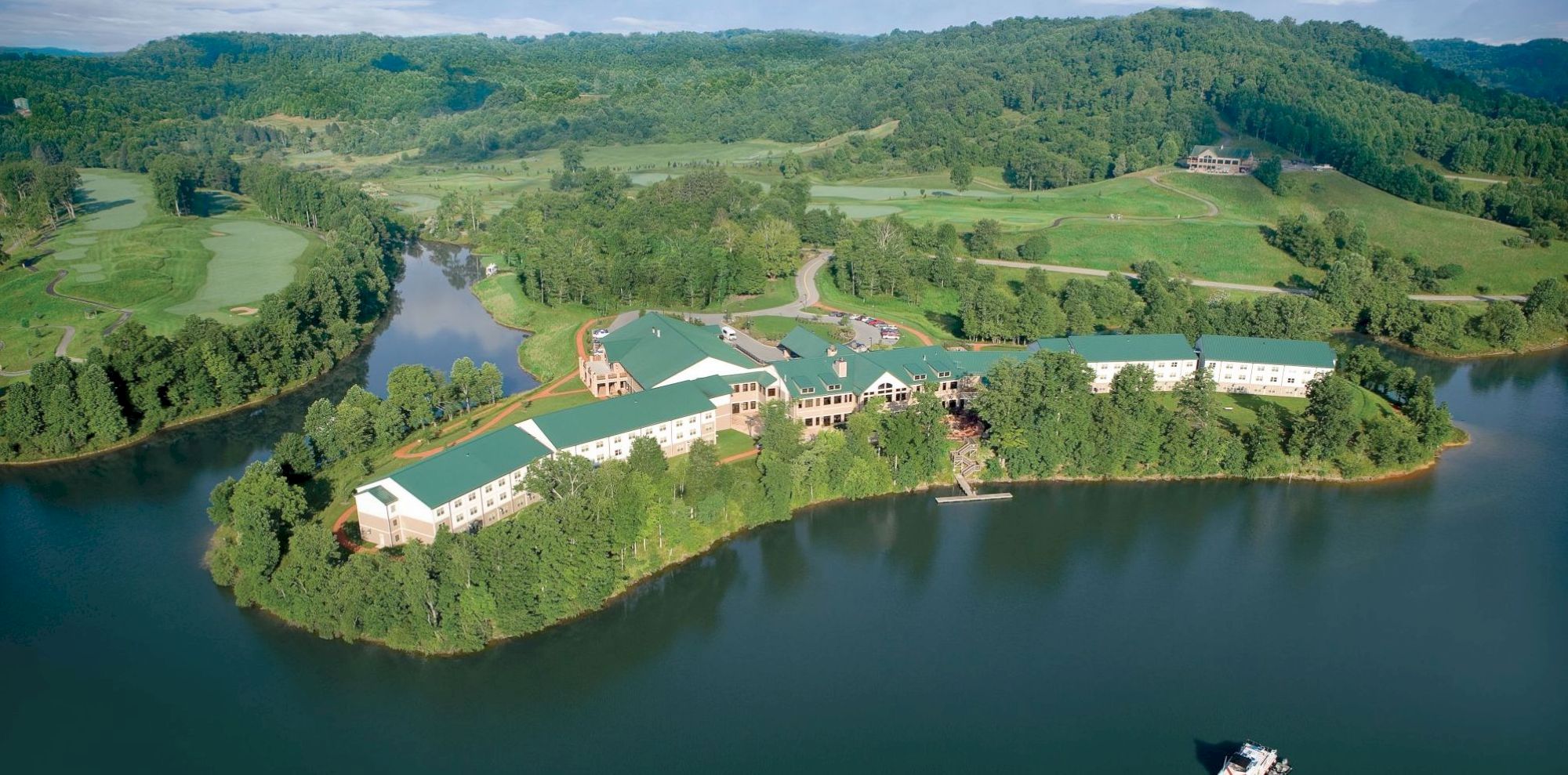 An aerial view of a large complex with green roofs situated on a wooded peninsula surrounded by a lake, with hilly terrain in the background.