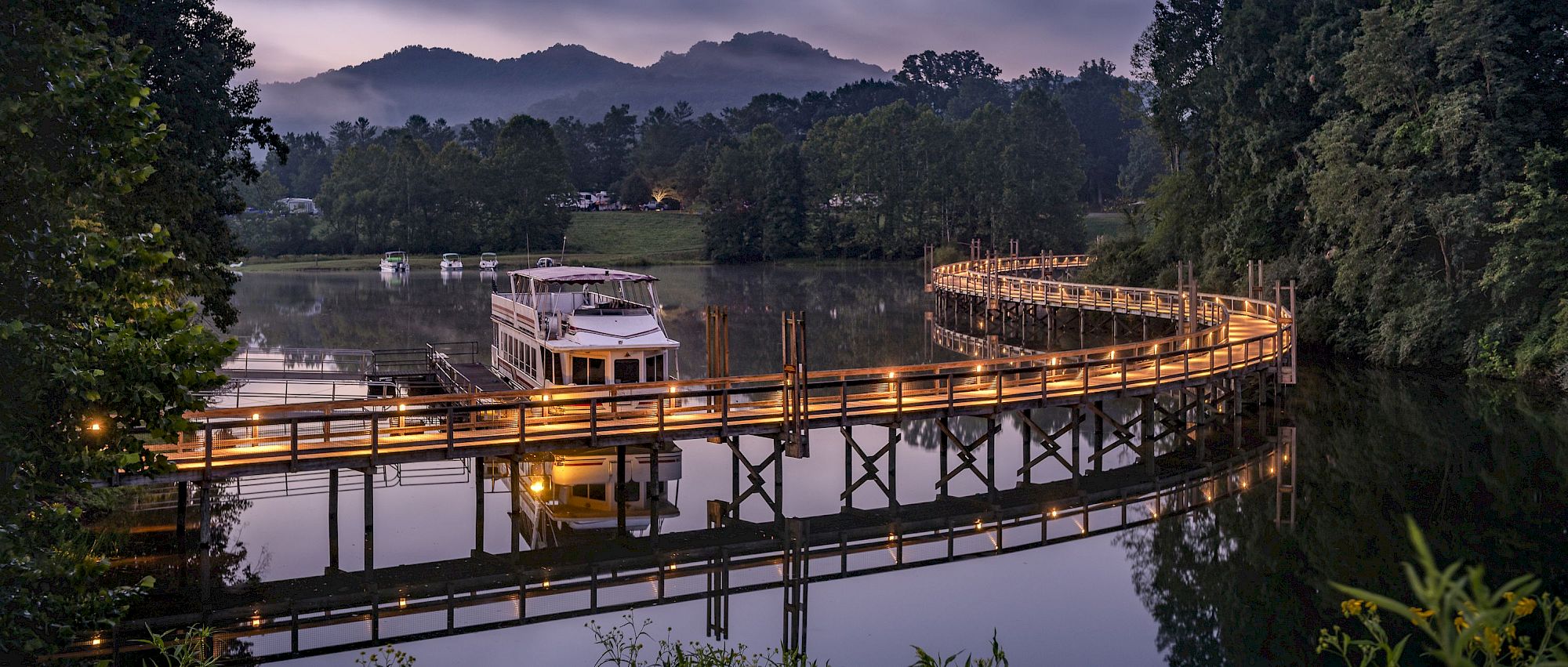 A boat on a serene lake is connected to a beautifully lit, curved wooden bridge, surrounded by lush greenery and distant mountains, ending the sentence.