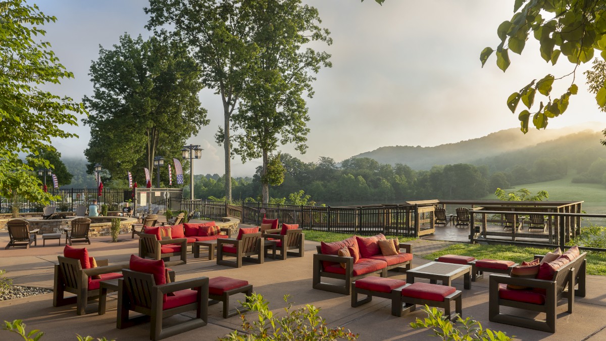 The image shows an outdoor seating area with red-cushioned chairs and tables, surrounded by greenery and overlooking hills in the background.