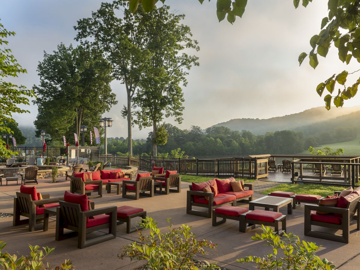 The image shows an outdoor seating area with red-cushioned chairs and tables, surrounded by greenery and overlooking hills in the background.