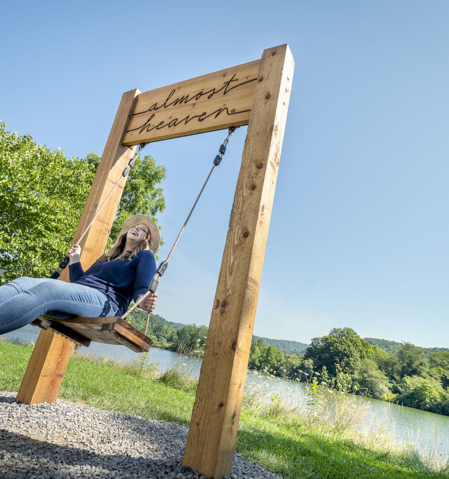 A person is swinging on a large wooden swing by a scenic lake with trees in the background. The structure has 