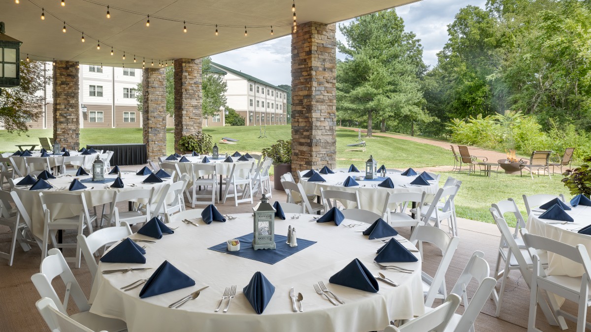 The image shows an outdoor event setup with round tables covered in white tablecloths, blue napkins, and white chairs, under string lights, ending the sentence.