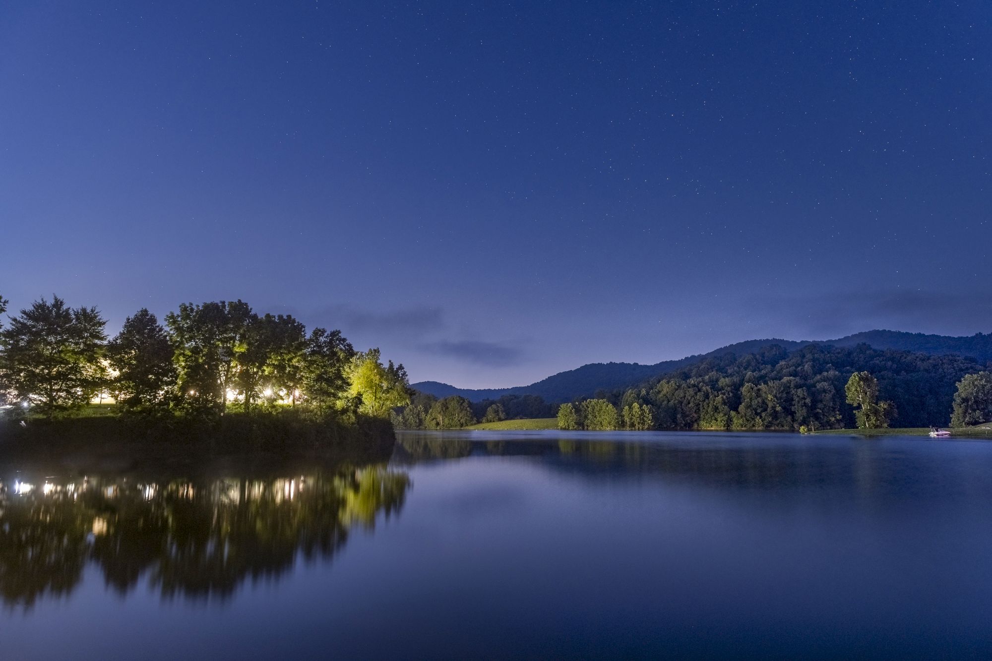 A serene lake scene at night, with lights illuminating trees on the left and a tranquil reflection on the water, under a clear, starry sky.