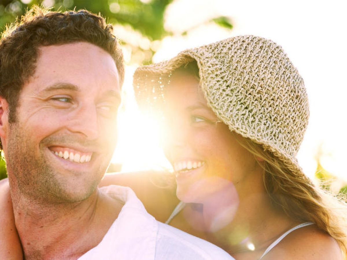 The image shows a couple smiling and looking at each other joyfully outdoors, with sunlight streaming from behind. Both appear happy and relaxed.