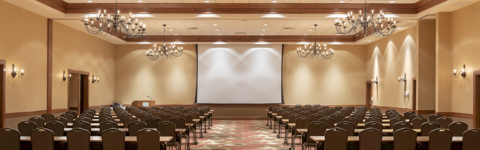 An empty conference room with rows of chairs, facing a large projection screen at the front, and ornate chandeliers hanging from the ceiling.