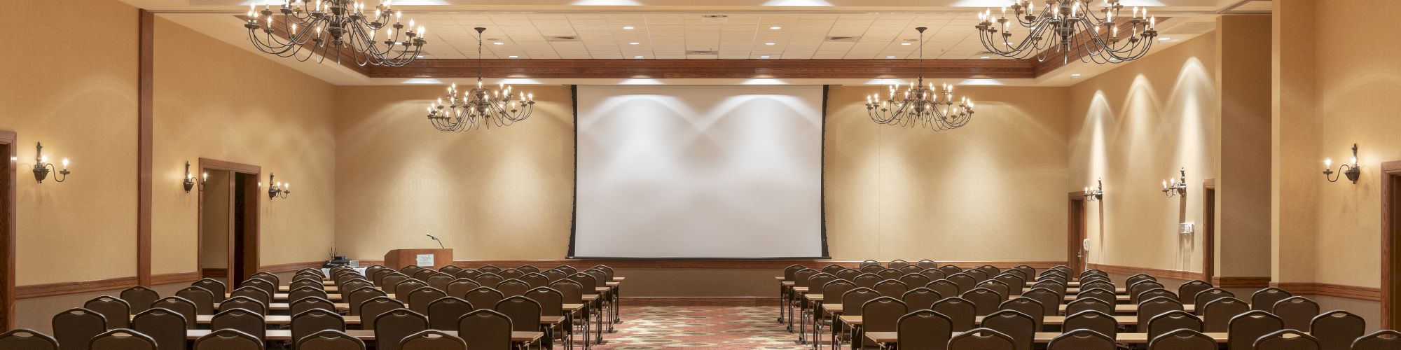 An empty conference room with rows of chairs, facing a large projection screen at the front, and ornate chandeliers hanging from the ceiling.