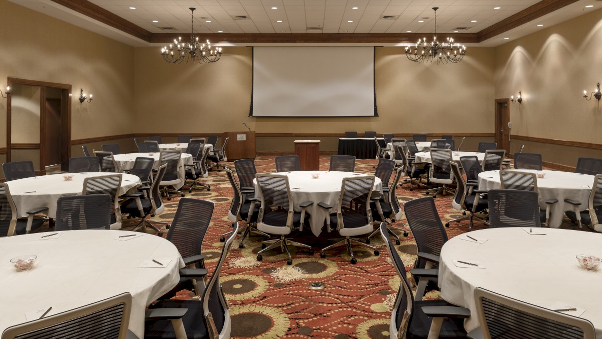 A conference room setup with round tables, chairs, a podium, and a projector screen in front, with a patterned carpet and chandeliers overhead.