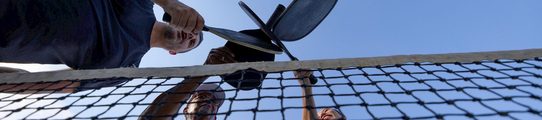 Four people are playing pickleball outdoors under a clear sky, using paddles and a ball on a net.