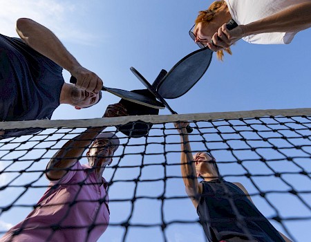 Four people are playing pickleball outdoors under a clear sky, using paddles and a ball on a net.