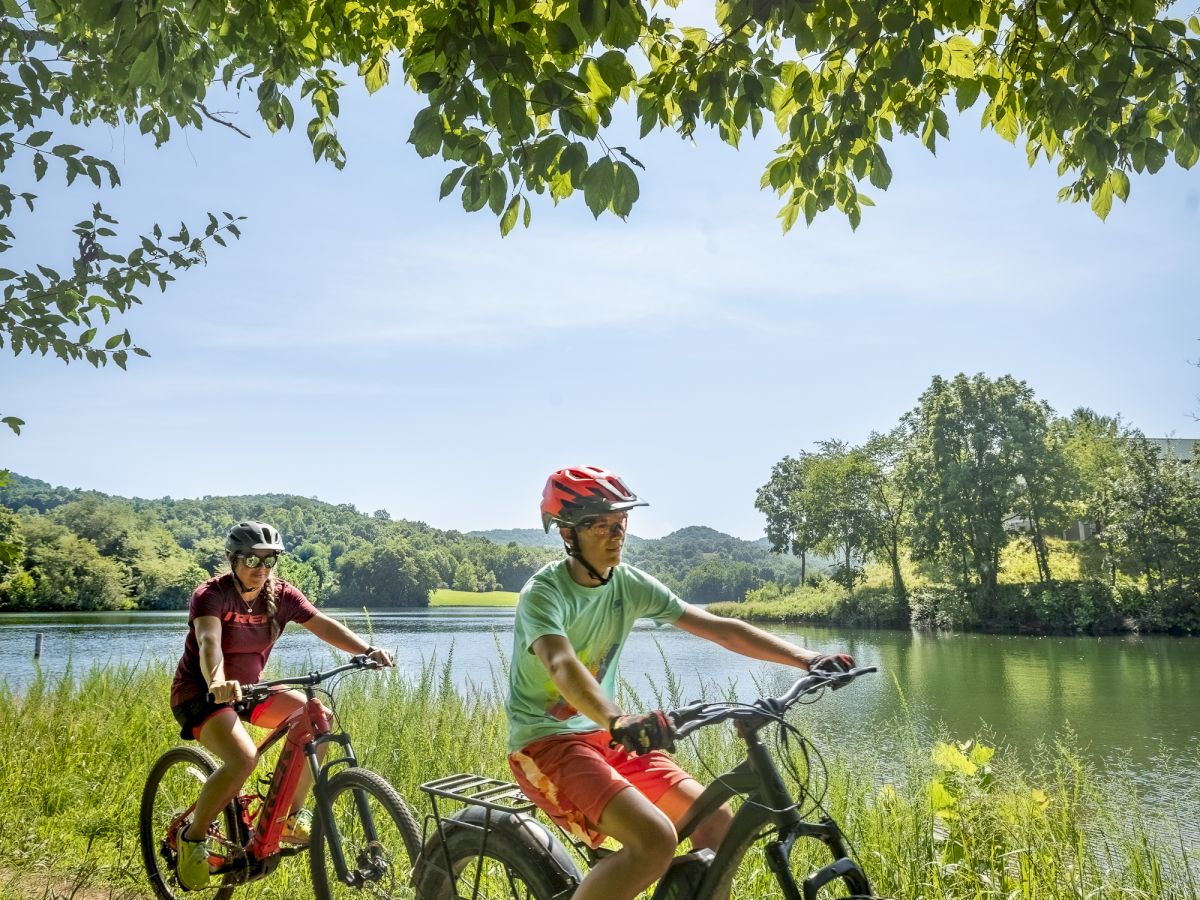 Two people are riding bicycles along a lakeside trail surrounded by trees and greenery under a sunny sky, with a shimmering lake in the background.