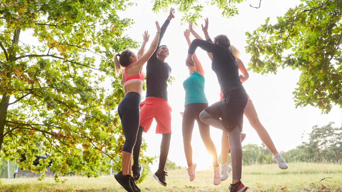 A group of five people in athletic wear are outside, jumping and reaching up, surrounded by trees and greenery, in a sunny setting.