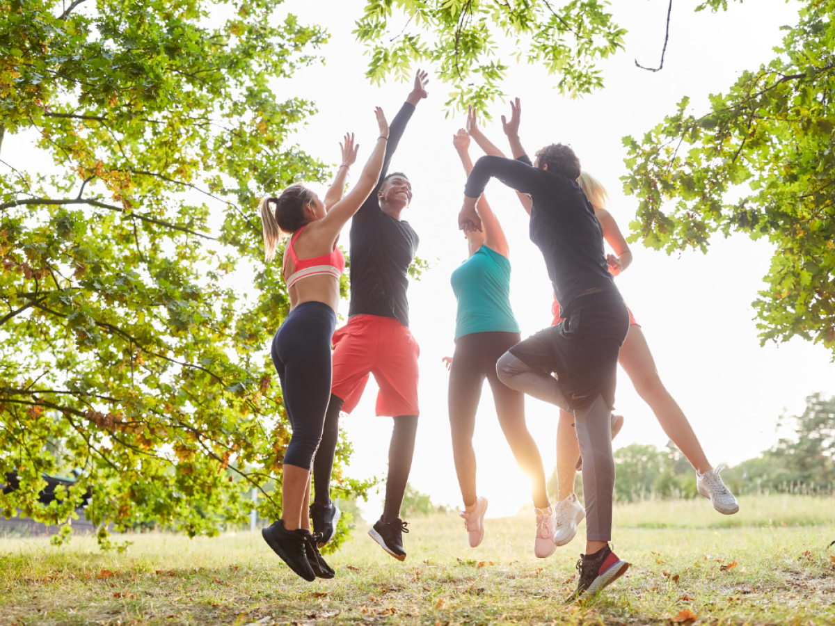 A group of five people in athletic wear are outside, jumping and reaching up, surrounded by trees and greenery, in a sunny setting.