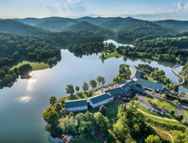 A large lake surrounded by green hills and trees, with a cluster of buildings and parking lot near the water's edge.