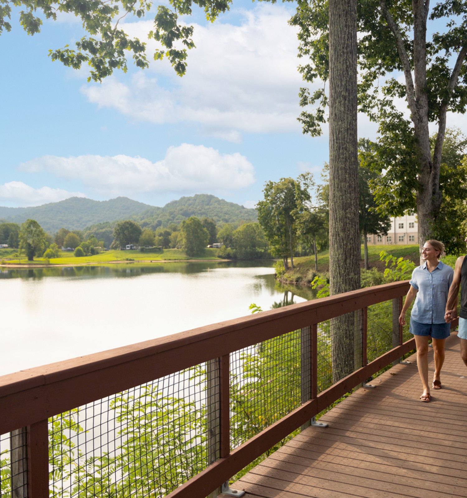 A couple walks hand-in-hand along a scenic, wooden lakeside boardwalk surrounded by trees and lush greenery on a sunny day.