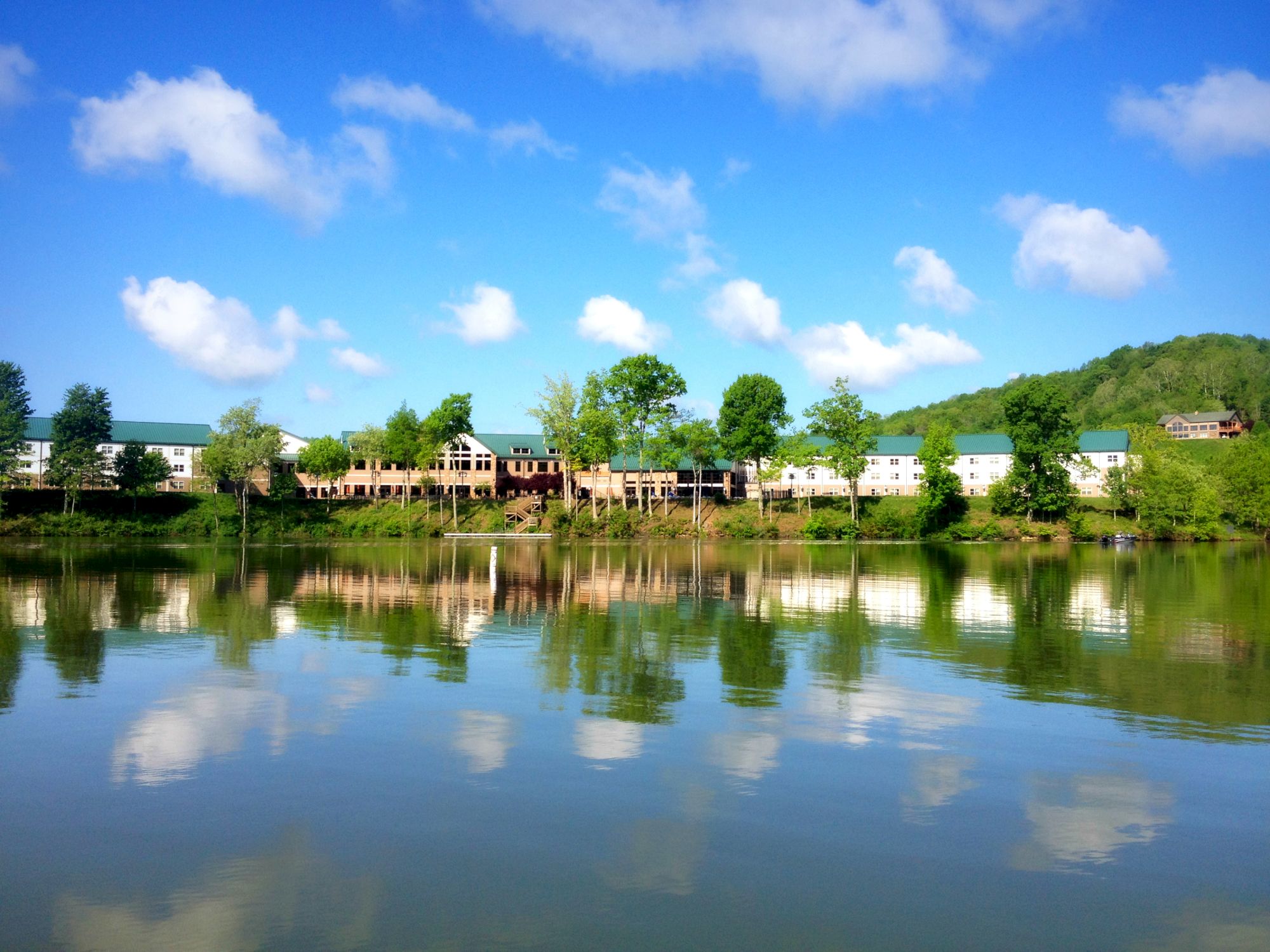 A scenic view of buildings and trees reflected in a calm body of water, under a bright blue sky with scattered clouds.