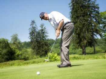A person is golfing, focusing on a putt on a green course with trees and blue sky in the background.
