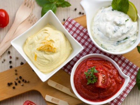 Three bowls of different sauces (mustard, white herb, and red tomato) on a wooden board with tomatoes, forks, and herbs around it.