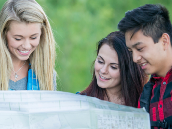 Three people are looking at a large map outside, smiling and appearing to enjoy themselves.