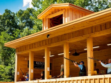 A wooden shooting range with people aiming rifles, set against a background of green trees and a blue sky, is shown in the image.