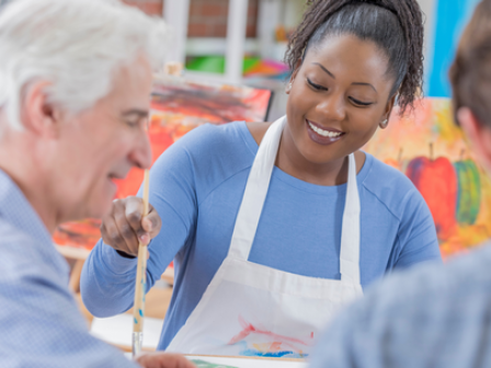 A woman assists two people with a painting project in a classroom setting, all smiling and wearing aprons.