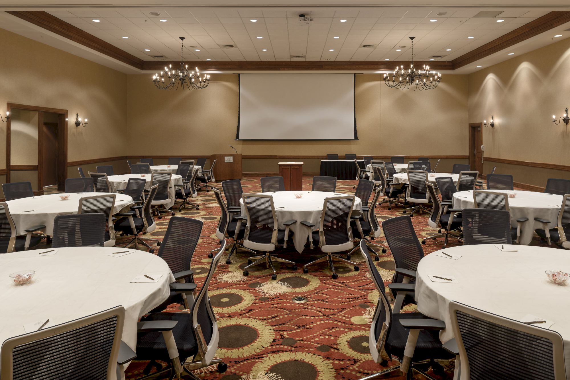 The image shows a conference room with round tables, chairs, chandeliers, and a projector screen at the front, set up for an event or meeting.