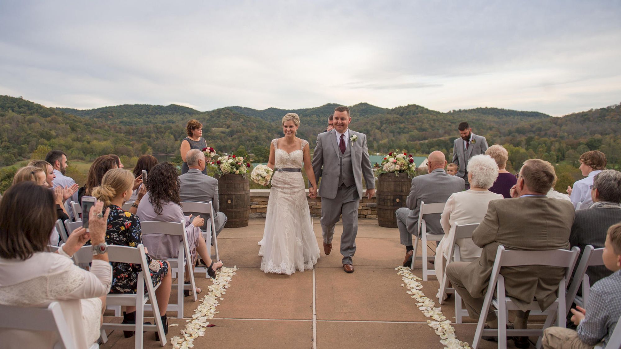 A bride and groom walk down the aisle at an outdoor wedding ceremony with mountains in the background and guests seated on either side.