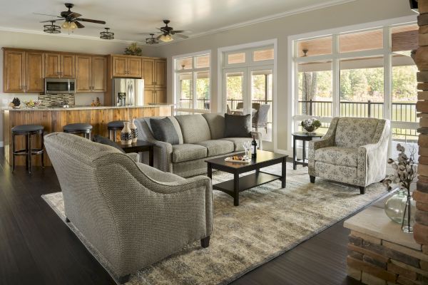 A cozy living room with grey furniture, a patterned armchair, and a kitchen. The room features large windows and a rug on a dark floor.