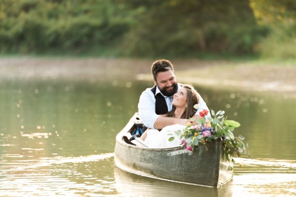 A couple in a canoe on a serene body of water, with the woman holding a bouquet of flowers and the man embracing her happily, ending the sentence.