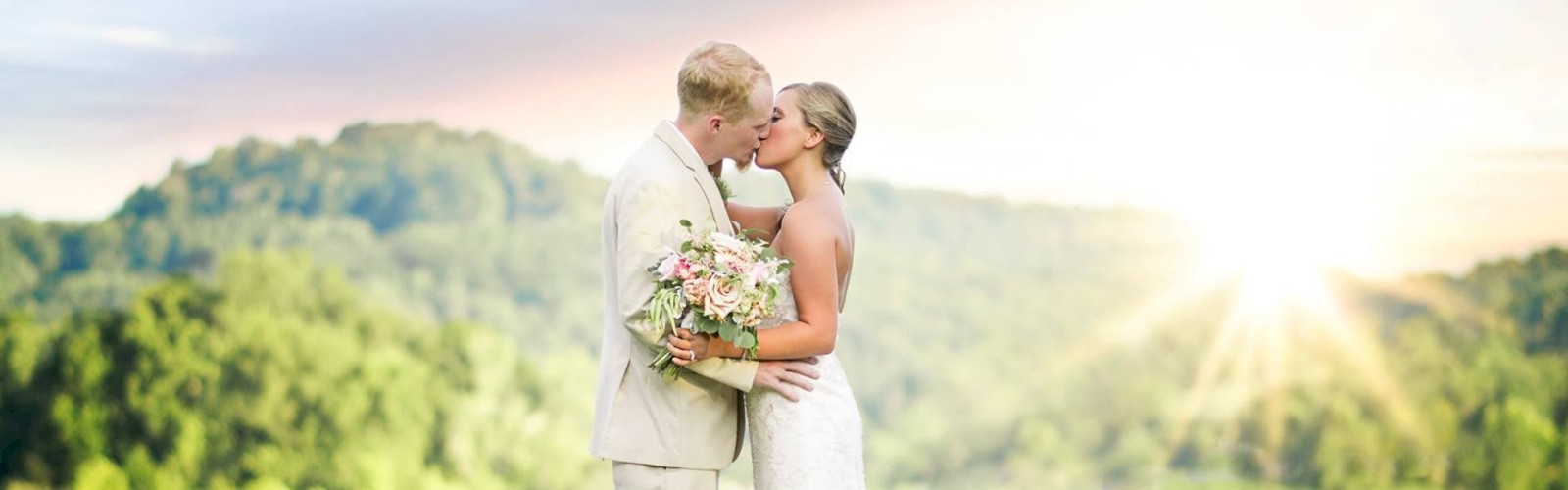 A couple kisses outdoors during their wedding, with the sun setting behind them over a scenic forest and lake.