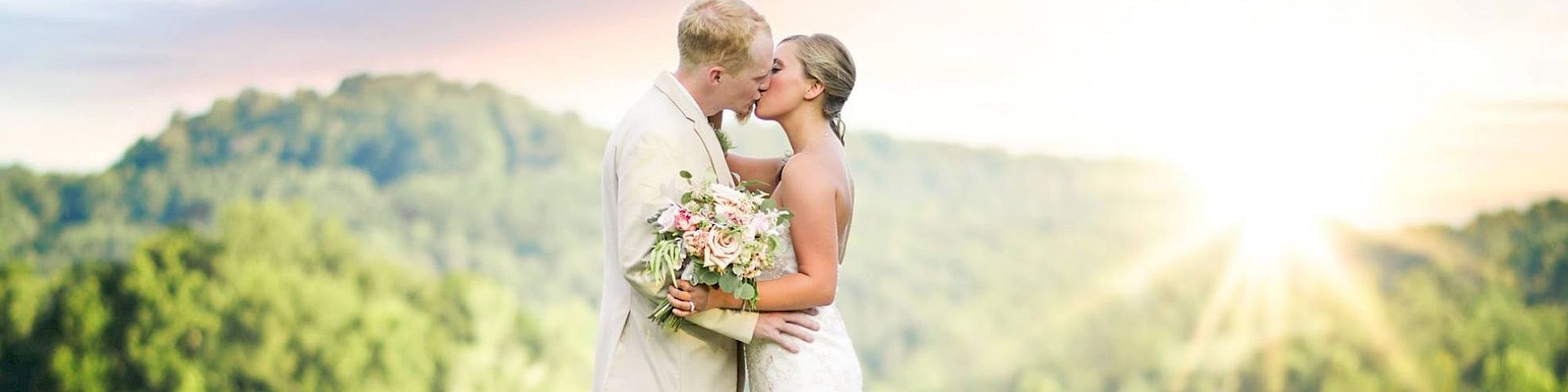 A couple kisses outdoors during their wedding, with the sun setting behind them over a scenic forest and lake.