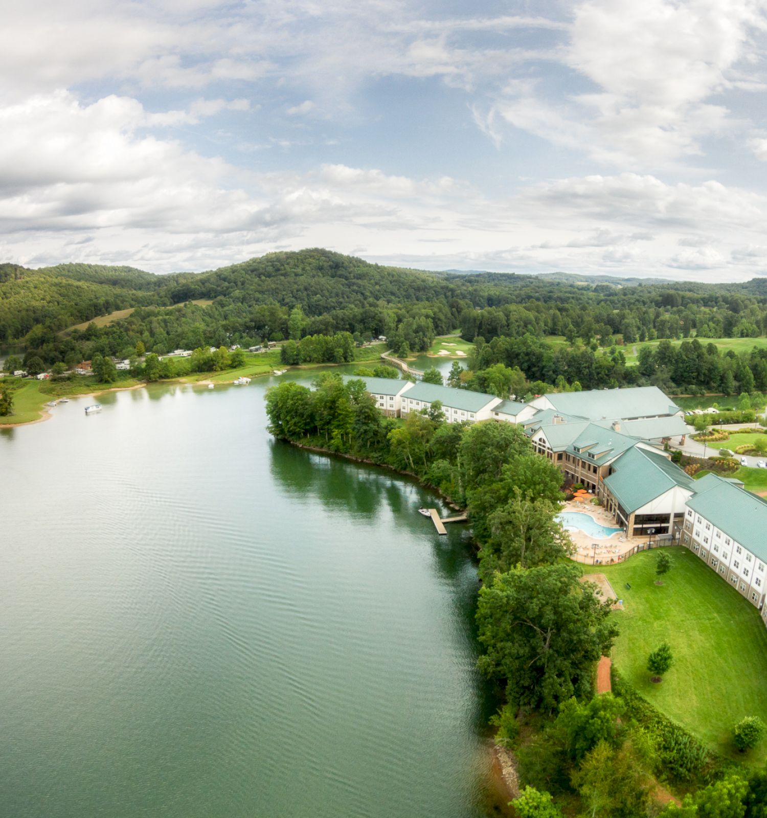 Aerial view of a large lakeside resort with green roofs, surrounded by lush forest and hills under a cloudy sky.