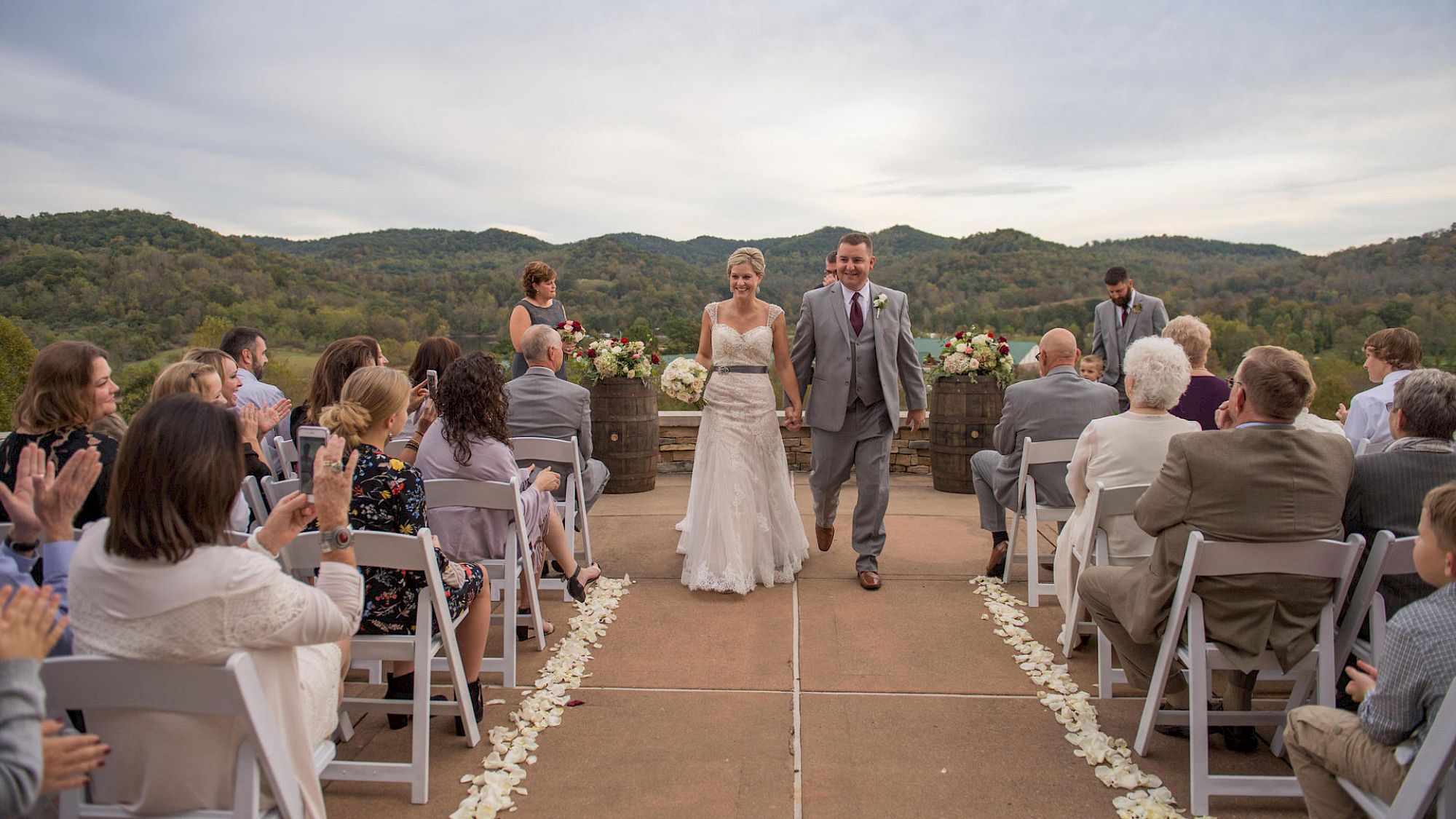 A bride and groom are walking down the aisle outdoors, surrounded by guests seated on both sides, against a scenic backdrop of hills and sky.