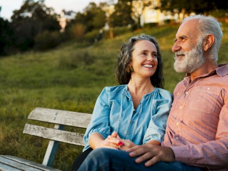 An elderly couple sits on a park bench, gazing at each other affectionately, enjoying a beautiful outdoor moment together.