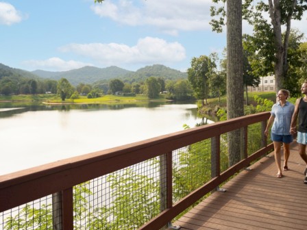 A couple is walking hand-in-hand on a scenic wooden trail by a peaceful lake with mountains in the background.