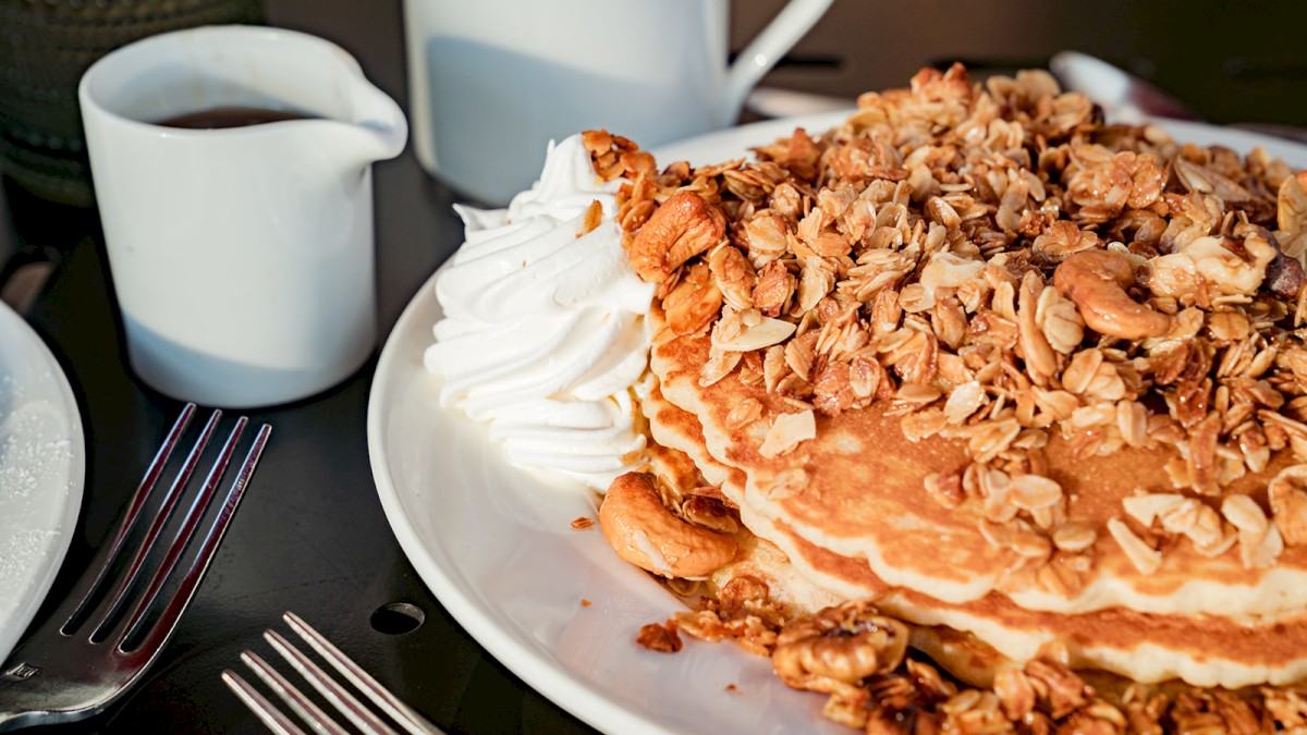 A plate of pancakes topped with granola and whipped cream, alongside a small pitcher of syrup and a couple of forks on the table.