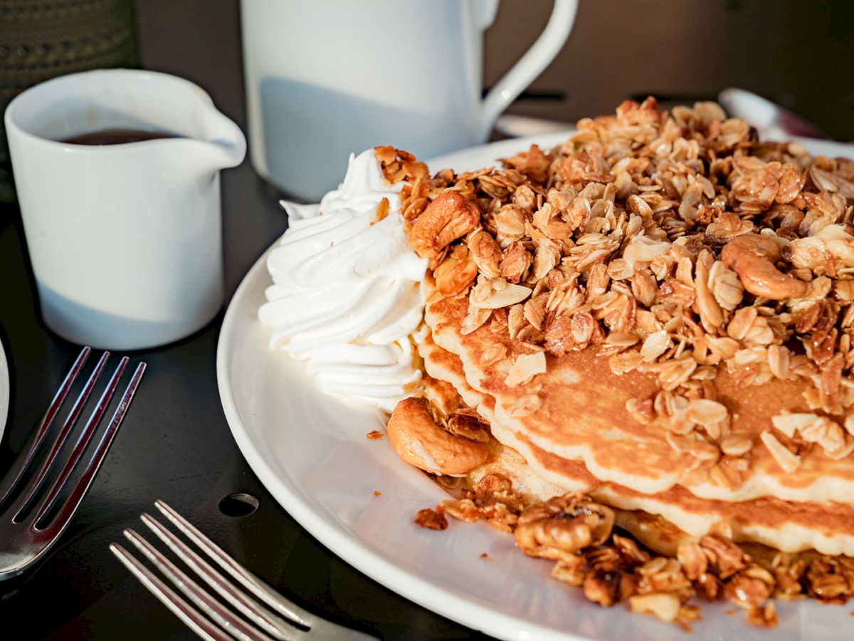 A plate of pancakes topped with granola and whipped cream, alongside a small pitcher of syrup and a couple of forks on the table.