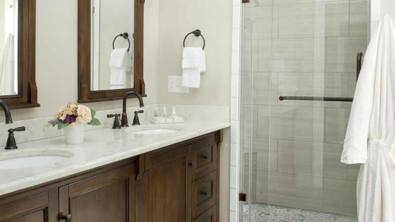 A modern bathroom with a wooden double vanity, marble countertop, wall-mounted lights, mirrors, and a glass-enclosed shower.