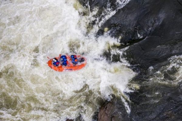 The image shows an aerial view of a group of people white-water rafting in a river with strong currents and rocks.