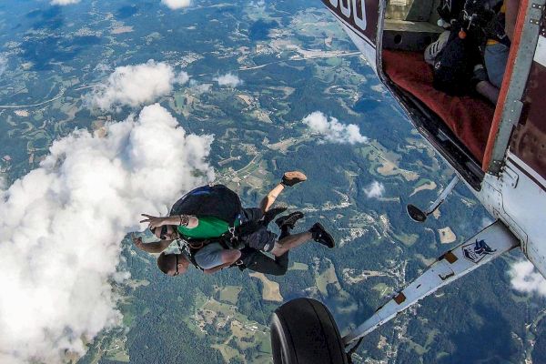 Two people are skydiving, having just jumped from a small aircraft. The view shows the airplane and vast landscape below.