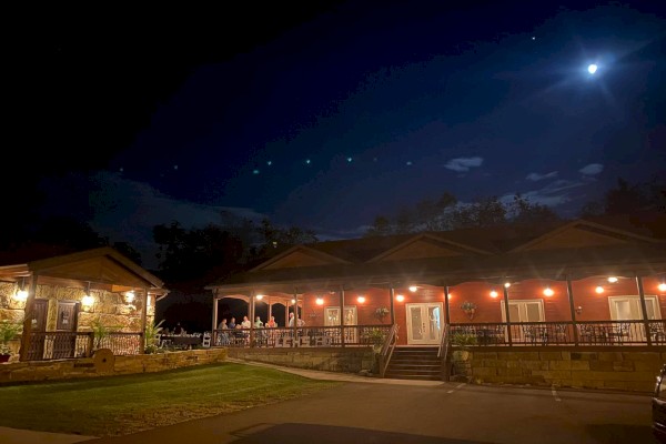An illuminated building stands under a night sky with a bright moon, featuring outdoor seating areas and warm lighting throughout the property.