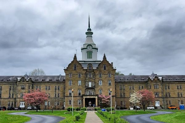 A historic, multi-story building with a tall spire, surrounded by green lawns, curved driveways, and spring trees, under a cloudy sky.