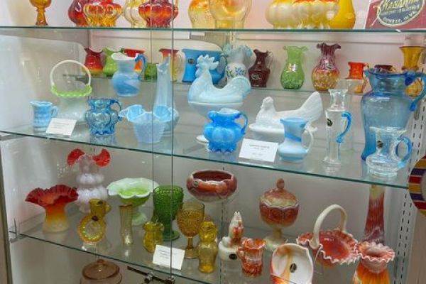 A display cabinet filled with various vintage glassware in bright colors, including pitchers, vases, and decorative items, arranged on shelves.