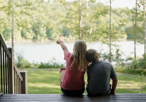 Two children sit on a porch, gazing at a peaceful lake surrounded by trees, with one child pointing out at the scenic view.