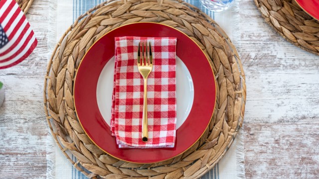 A red-and-white checked napkin with a gold fork is placed on a white plate, set on a red-rimmed charger, with woven placemats and a rustic wooden table.