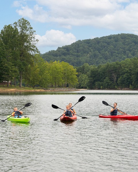 Three people are kayaking on a calm lake with lush trees and hills in the background on a partly cloudy day.