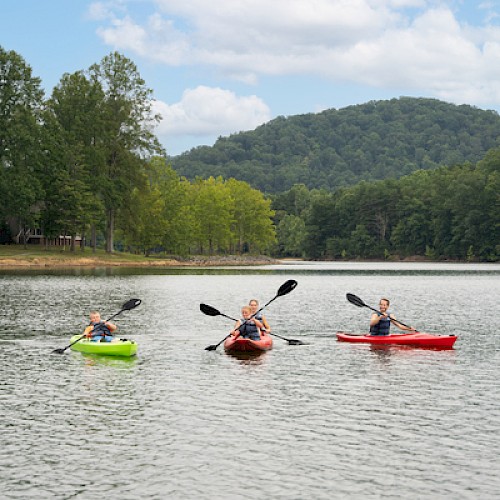 Three people are kayaking on a calm lake with lush trees and hills in the background on a partly cloudy day.